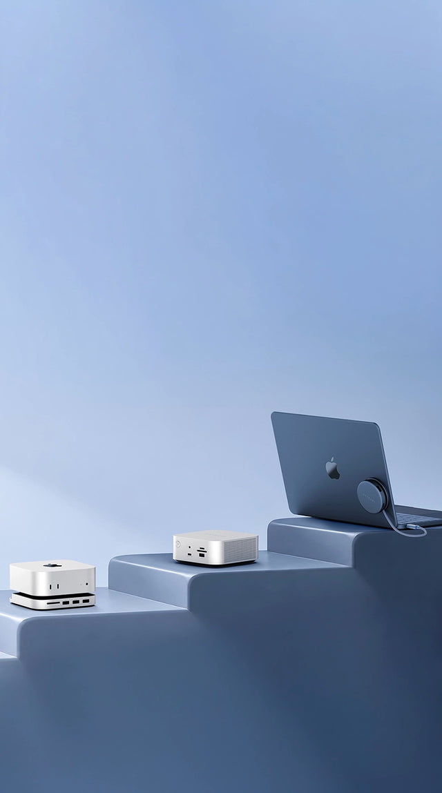 Satechi Apple accessories displayed on light blue steps, left to right:  a silver Mac mini sitting on the Mac Mini M6/M5 Pro/M4 Stand & Hub with SSD Enclosure, with an SD card slot and USB ports on the front; the silver Thunderbolt 5 CubeDock with SSD Enclosure, with front ports and a power button; and the round OntheGo 5-in-1 Multiport Adapter mounted on MacBook Neo.