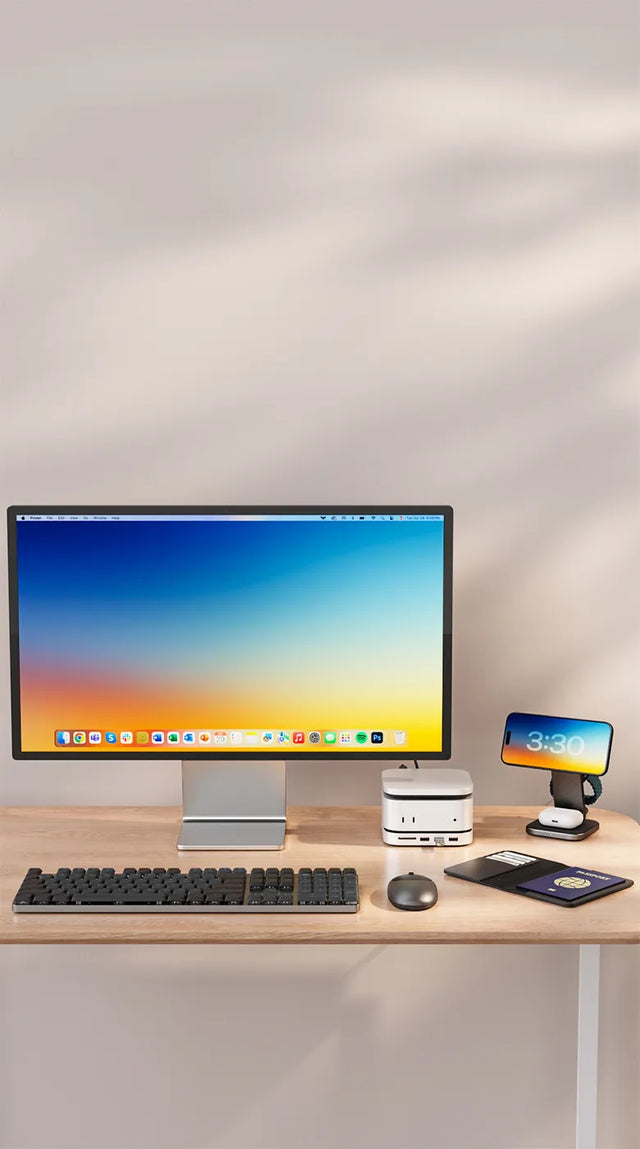 A modern desktop setup featuring a large monitor on a wooden desk with the Satechi Mac Mini M4 Stand & Hub with SSD Enclosure placed beneath the display, alongside the Satechi USB4 Slim NVMe SSD Enclosure. In front of the monitor sits the Satechi SM3 Slim Mechanical Backlit Bluetooth Keyboard and the Satechi M1 Bluetooth Mouse. To the right, a smartphone rests on the Satechi 3‑in‑1 Foldable Qi2 Wireless Charging Stand, next to a Vegan‑Leather FindAll Passport Cover.