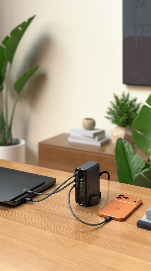 Vertical view of a wood desk with the Satechi ChargeView™ 240W GaN charger upright in its base, three gray braided USB-C cables connecting a space black iPad Pro to the left and an orange iPhone to the right. The real-time LCD reads: C1 100 W; C2 35 W; C3 30 W; C4 – – W; C5 – – W; C6 – – W. Behind it, a cream wall with framed abstract art, tall green plants, and a wood credenza holding a stack of white books and a round ceramic vase.