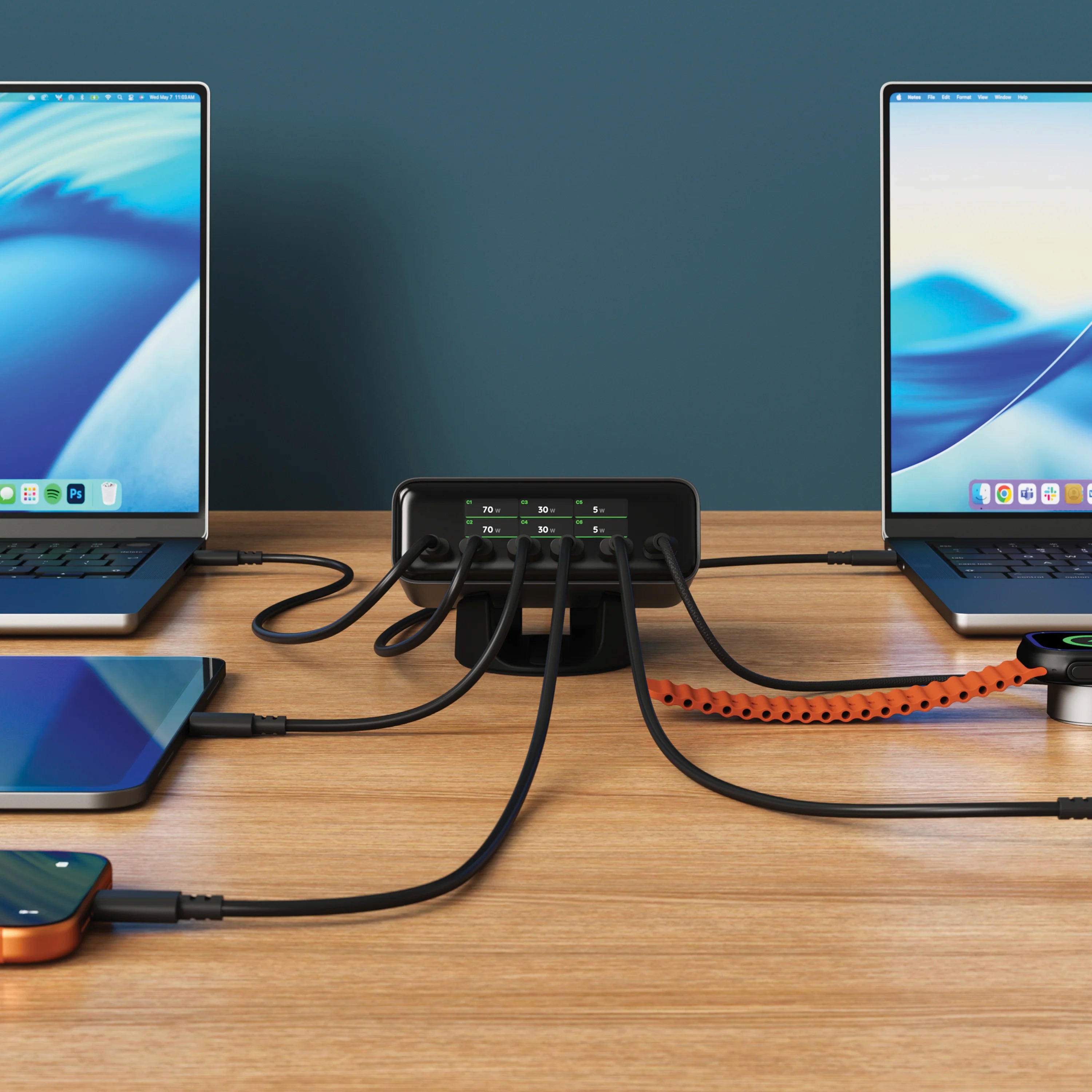 Satechi ChargeView™ 240W Desktop Charger in black positioned horizontally on a wood desk against a dark teal wall, acting as a charging station for multiple devices with six USB-C cables fanning out to a MacBook on the left, a MacBook on the right, a tablet, an orange iPhone, and an Apple Watch charging puck connected by an orange braided cable. The front-facing display of the 6-port USB-C charger reads: C1 70 W; C2 70 W; C3 30 W; C4 30 W; C5 5 W; C6 5 W. The Apple Watch screen shows a green charging bolt