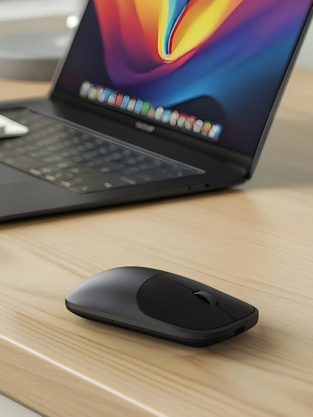 A closeup of the Slim EX Wireless Mouse next to a MacBook on a light wood table.