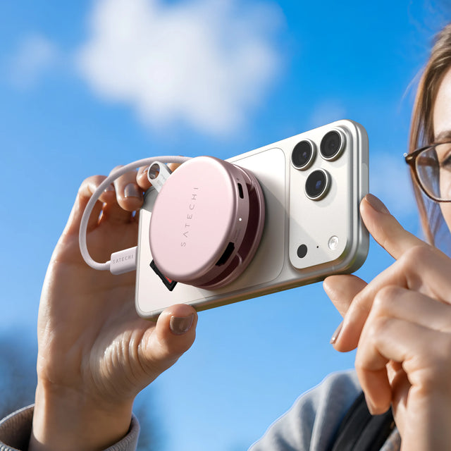 A person's hands hold a white iPhone against a bright blue sky, with a Blush Satechi magnetic USB-C hub attached to the back via MagSafe, its coiled braided cable and a USB flash drive visible. Headline reads "Magnetic mounting to your Neo's lid or MagSafe iPhone. The hub that moves the way you do." Port label "HDMI 4K/60Hz" visible on the hub.