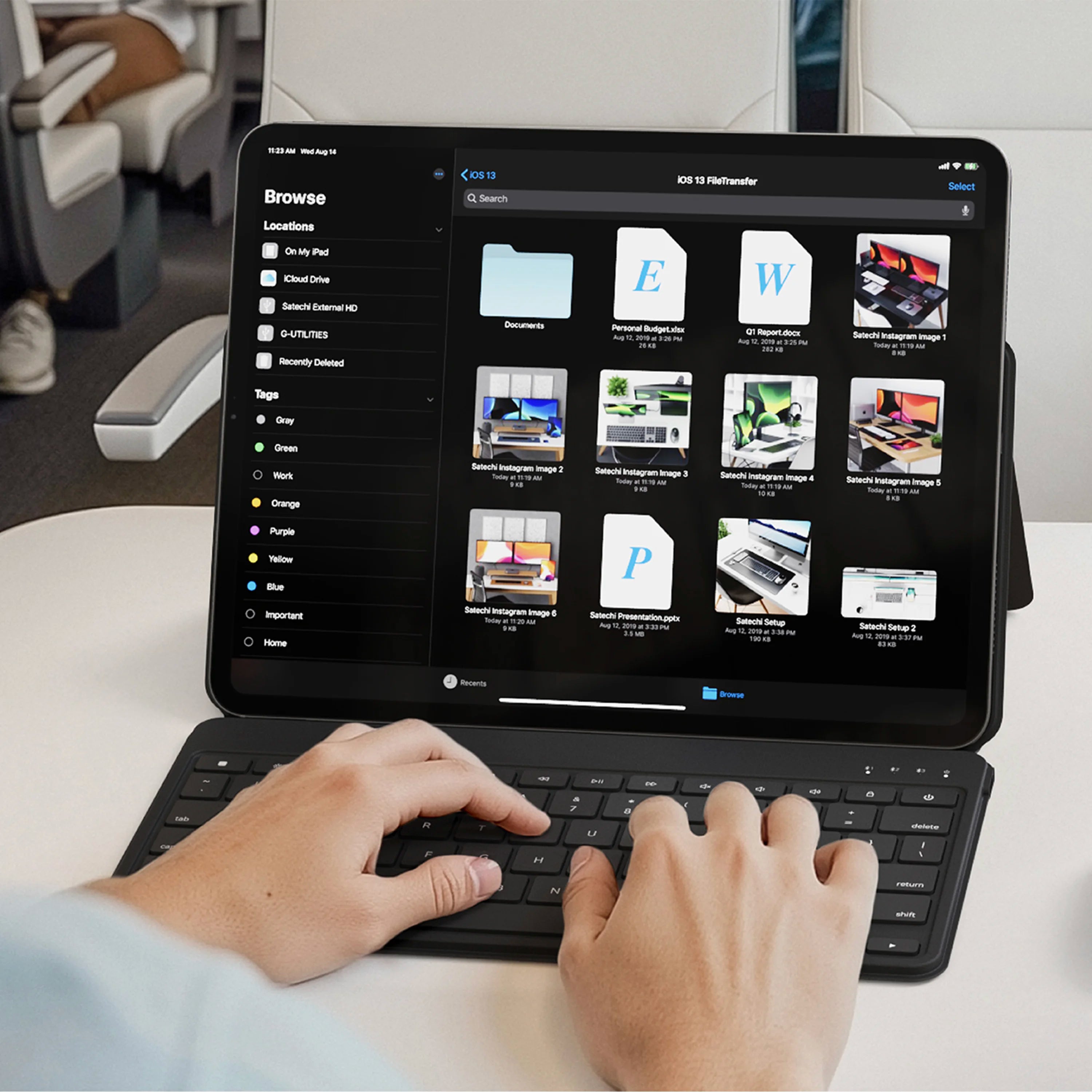 Satechi OnTheGo™ Bluetooth Keyboard with Stand in black, a compact Bluetooth keyboard for iPad shown in use on a white tray table aboard a train or aircraft cabin, with a person's hands typing on the slim keys and the protective cover folded behind the tablet as an adjustable stand. The keyboard's top row shows media and function keys including rewind, play/pause, fast-forward, volume down, mute, volume up, screenshot and power icons, with tab, caps, delete, return and shift keys visible. The iPad screen di