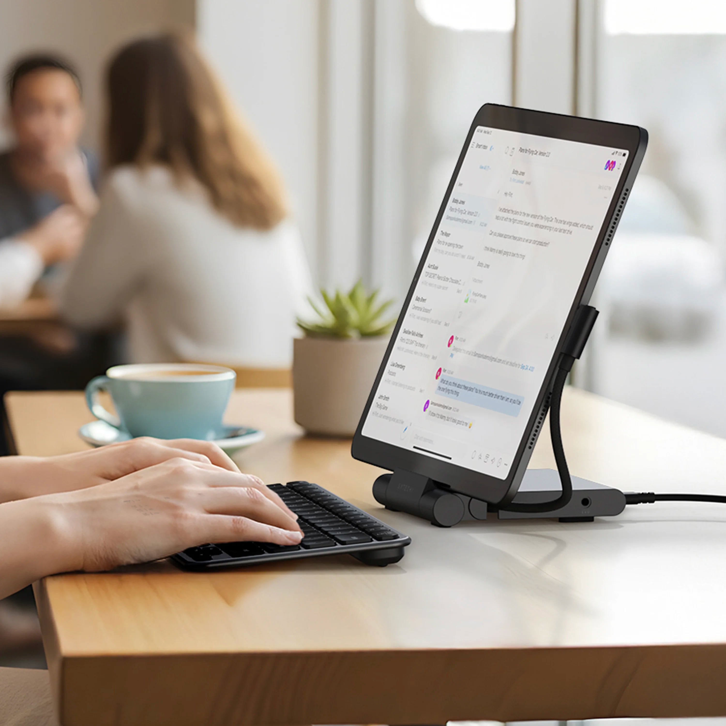 Satechi OnTheGo™ Foldable Stand Hub in space gray holding an iPad upright in portrait orientation on a light wood café table, functioning as a foldable iPad stand and USB-C docking station in one. The hub's hinged base is folded out flat with the aluminum arm supporting the tablet from behind, a black USB-C cable plugged into the side and a 3.5mm headphone jack visible on the right edge. A woman's hands type on a slim black wireless keyboard in the foreground, with a light blue cup of coffee on a saucer and