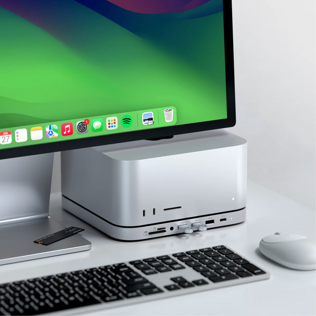 A silver Mac Studio sits on the Satechi Stand & Hub For Mac Studio M5 With NVMe SSD Enclosure, a Mac Studio stand with front ports, next to a monitor, keyboard, and white mouse on a white desk. Two USB flash drives are plugged into the hub, and a WD_BLACK SN850X NVMe SSD rests on the monitor stand.