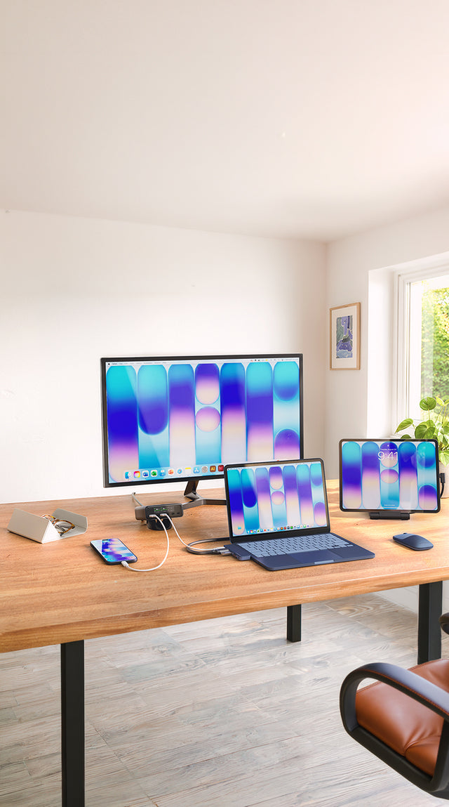 An Indigo MacBook Neo on a light wood desk with an Indigo Satechi USB-C Snap Hub plugged directly into the laptop's side ports, sitting flush against the chassis and running a cable out to a large external monitor. A Satechi ChargeView 240W Desktop Charger sits behind the monitor stand, it displays showing live per-port wattage as it powers the MacBook through the USB-C hub and charges an iPhone on the desk. An iPad stands upright in a Satechi OnTheGo Foldable Stand Hub, its lock screen reading "9:41".