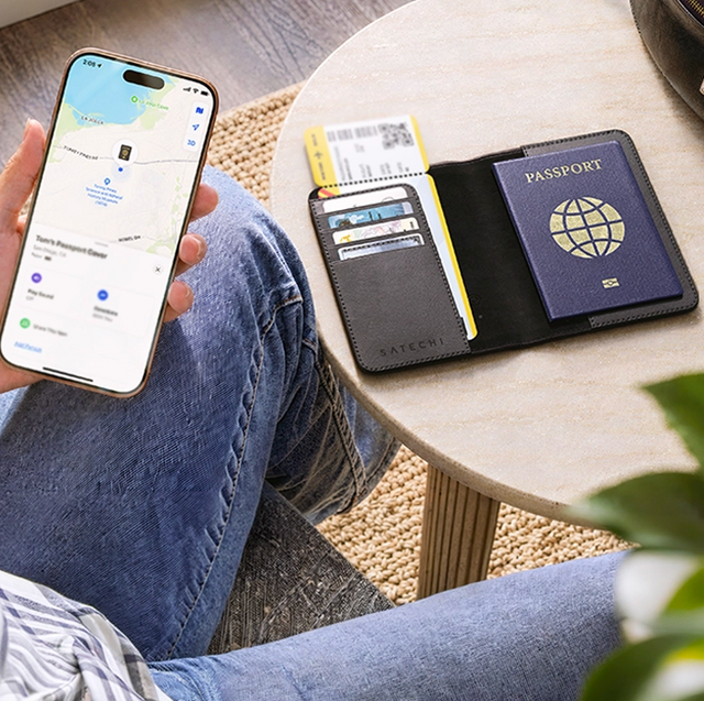 Satechi FindAll Passport Cover with Find My tracking on a table holding a passport, cards and a boarding pass, while a person checks its location in the Find My app on an iPhone.