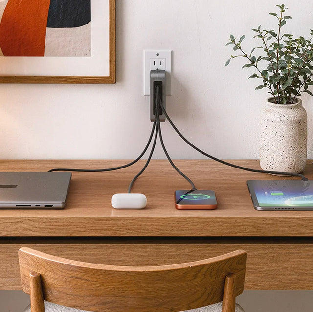 Satechi 145W USB-C 4-Port GaN Travel Charger plugged into an outlet above a desk, charging a laptop, AirPods case, phone on a wireless pad, and an iPad through three cables.