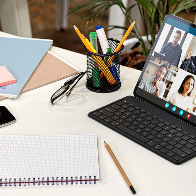 A modern home office workspace with a tablet displaying a virtual meeting, positioned on a white desk, on top of a Satechi OntheGo™ Bluetooth Keyboard with Stand. Nearby is a notebook, pencil, smartphone, folders, and a cup filled with colorful pens and markers.