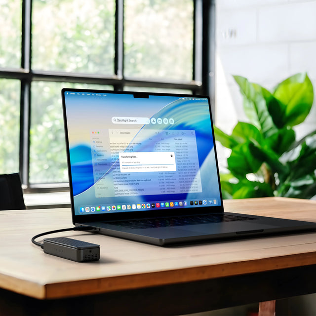 Close-up detail of the black anodized DotDisk Thunderbolt 5 SSD enclosure on the left. On the right, a MacBook Pro on a wood desk with the silver DotDisk connected, showing a Finder window with a "Transferring files..." dialog at 42% complete.