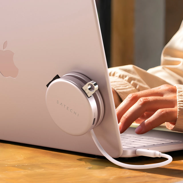 Rear view of a blush MacBook Neo with a color-matched blush OntheGo 5-in-1 multiport adapter to the lid and a coiled USB-C cable, hands typing while wearing a cozy cream sweater, showing student uploading files from SD card and flash drive. 