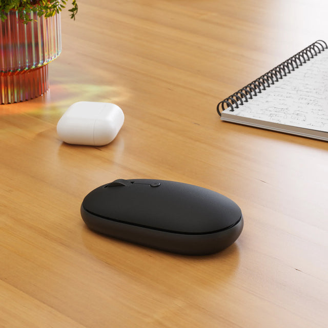 Black OntheGo™ Bluetooth Mouse on a wooden desk with a white Airpod case and notebook in the background.