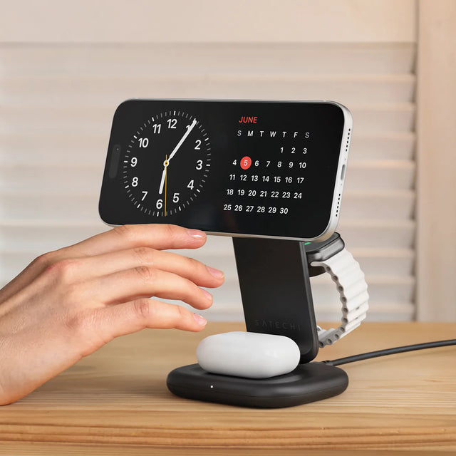 A hand reaching out to adjust a Satechi 3-in-1 Foldable Wireless Charging Stand with Qi2 25W on a nightstand, charging an iPhone, Apple Watch, and AirPods.