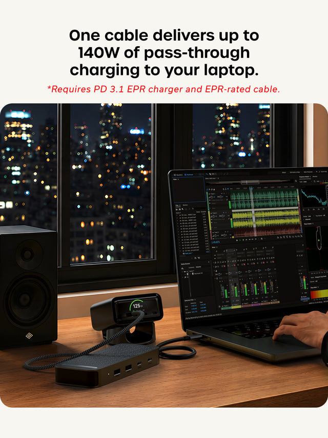 One cable delivers up to 140W of pass-through charging to your laptop. *Requires PD 3.1 EPR charger and EPR-rated cable. The Satechi ExtendView USB4 Dual HDMI Multiport Adapter sits on a wood desk at night in front of a window with city lights. A braided cable runs from the adapter to a desktop charger whose screen shows 125W, and the adapter’s host cable plugs into a laptop running multitrack audio editing software. A hand rests on the trackpad, and a bookshelf speaker stands to the left.