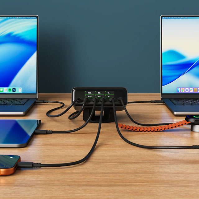 Satechi ChargeView™ 240W Desktop Charger in black positioned horizontally on a wood desk against a dark teal wall, acting as a charging station for multiple devices with six USB-C cables fanning out to a MacBook on the left, a MacBook on the right, a tablet, an orange iPhone, and an Apple Watch charging puck connected by an orange braided cable. The front-facing display of the 6-port USB-C charger reads: C1 100 W; C2 100 W; C3 30 W; C4 30 W; C5 5 W; C6 5 W. The Apple Watch screen shows a green charging bolt