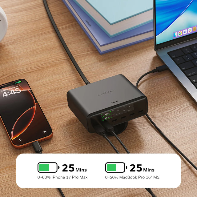 Satechi ChargeView™ 240W Desktop Charger in black lying flat on a wood desk beside an orange iPhone with a MacBook Pro to the right and a stack of  books behind. The SATECHI logo is  printed on the top surface, and the display on this 140W USB-C charger reads: C1 140 W with a green battery icon; C2 40 W; C3 – – W; C4 – – W; C5 – – W; C6 – – W.  Two green battery icons with the text: "25 Mins — 0~60% iPhone 17 Pro Max" and "25 Mins — 0~50% MacBook Pro 16" M5".