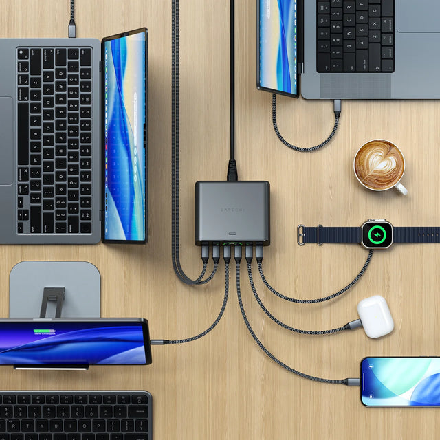 Overhead view of the Satechi ChargeView™ 240W Desktop Charger in space gray on a light wood desk, used as a MacBook charging station with six gray braided USB-C cables running from its front ports to a laptop and tablet at upper left, a MacBook Pro at upper right, an iPad on a stand at lower left, an AirPods case, an Apple Watch, and an iPhone at lower right. The SATECHI logo is centered on the top of the charging hub, a black power cable exits the rear, and a small green readout glows between the ports.