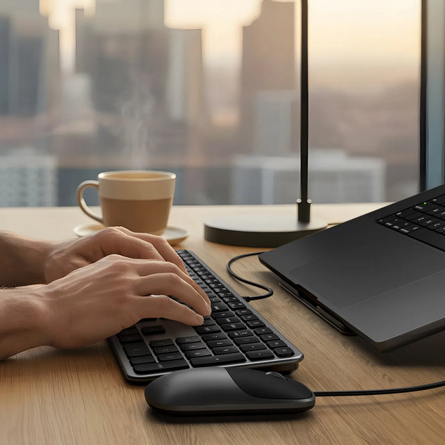 Person using a laptop with Slim LX3 Wired Keyboard and Slim LX Wired Mouse on a desk, with a cup of coffee and lamp in the background.
