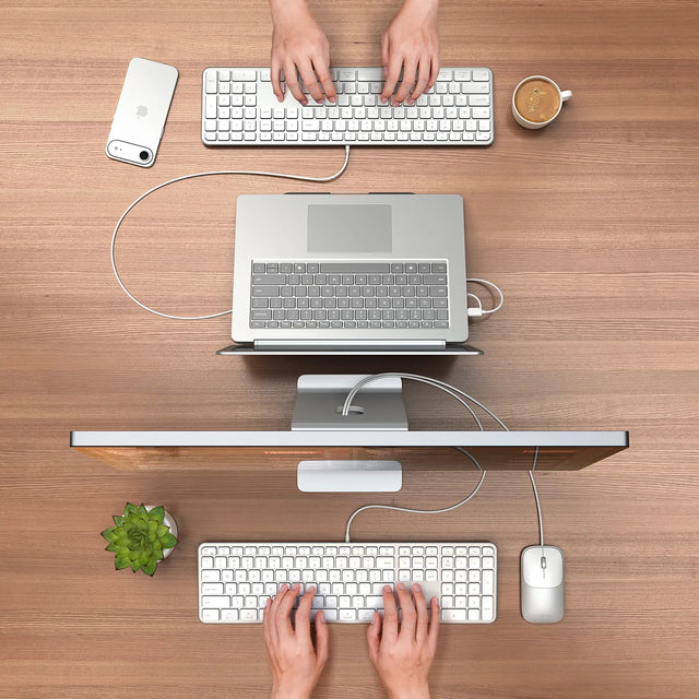 Two people typing on two Slim LX3 Wired Keyboards and two Slim LX Wired Mice, one tconnected to a MacBook and the other an iMac, on a wooden desk with a cup of coffee and an iPhone.