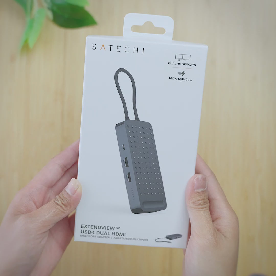 Unboxing video of the Satechi ExtendView™ USB4 Dual HDMI Multiport Adapter. Hands open the retail box, lift out the Space Black adapter and turn it to show the USB-C charging port, two HDMI ports, and two USB-A and two USB-C data ports. The adapter is then connected to a desk setup, plugging into a laptop and two external monitors.