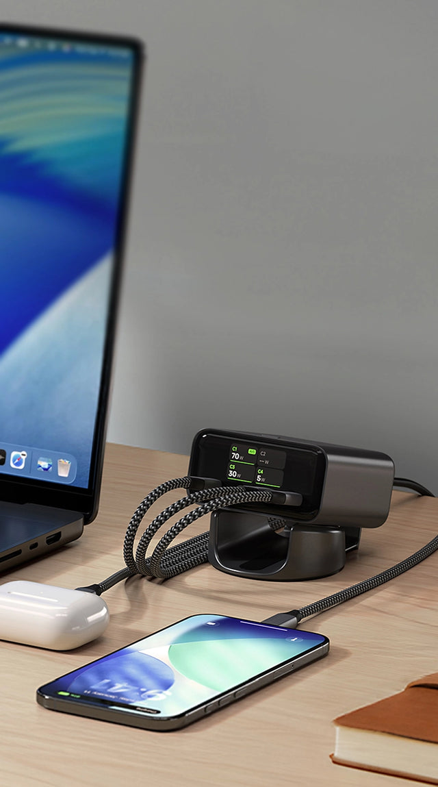 Satechi ChargeView™ 140W Desktop Charger on a wooden desk next to a MacBook, iPhone, and AirPods. The charger is positioned in a stand and connected via braided USB‑C cables. The front‑facing LCD display shows power distribution with illuminated green indicators, demonstrating real‑time power output while charging multiple devices on a desktop workspace.